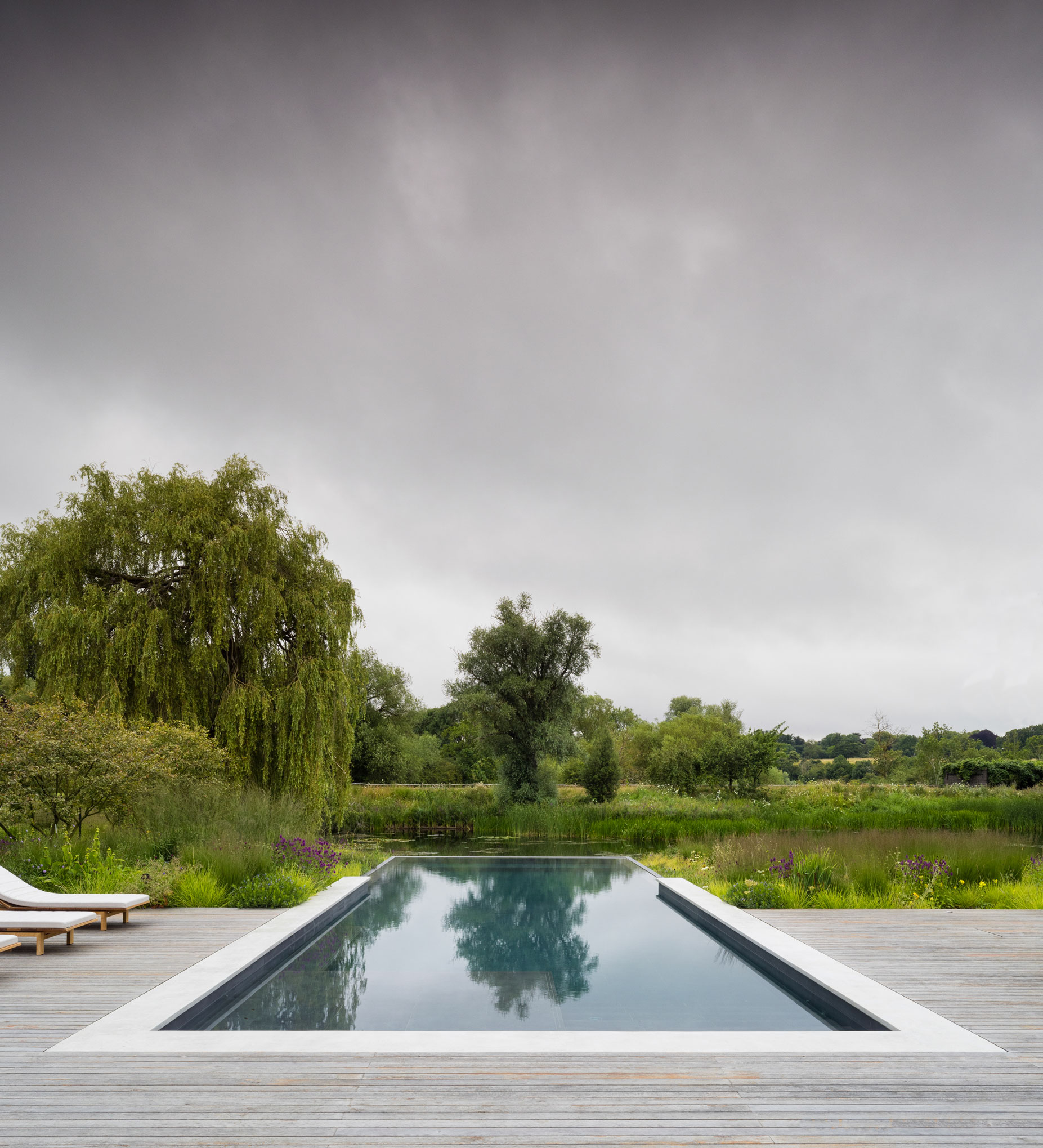 Colm Joseph modern country garden cambridgeshire swimming pool infinity edge timber deck sun loungers naturalistic planting design landscape view willow trees dramatic sky